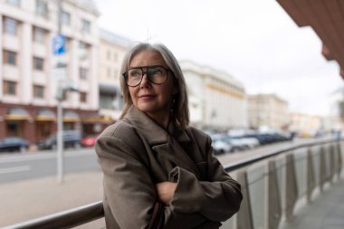 Orta yaşlı bir iş kadını hareketli bir şehir caddesinde duruyor, kollarını kavuşturup düşünceli bir şekilde uzaklara bakıyor, kariyerini ve planlarını serin öğleden sonra havasında yansıtıyor..