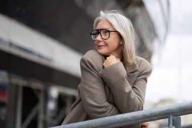 Yaşlı bir iş kadını, düşüncelere dalmış, korkuluklara yaslanıyor. Gözlük takıyor ve şık bir ceket giyiyor, hareketli bir şehir ortamında bilgelik ve deneyim duygusu taşıyor..