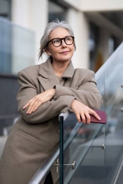 Yaşlı bir iş kadını, güvenli bir şekilde ofisinin dışında duruyor, kollarını parlak bir cama dayayıp, etrafını dikkatli bir ifadeyle seyrediyor..