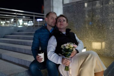 Bir adam ve bir kadın dışarıdaki taş merdivenlerde oturuyorlar. Kadın elinde bir buket çiçek tutuyor ve adam elinde bir kupa tutuyor. İkisi de yukarı bakıyor..