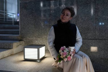 A woman in a white shirt and black vest is standing on concrete steps outside a building at night. She is holding a bouquet of pink flowers and a cup in her right hand. clipart