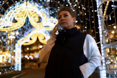 Siyah yelek ve beyaz süveter giyen bir kadın, parlak ip ışıklarıyla aydınlatılmış bir şekilde geceleri şehir caddesinde yürüyor. Telefonla konuşuyor, kameraya bakmıyor..