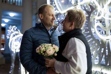 A man and woman walk through a city at night, illuminated by streetlights and decorations. The man is wearing a blue jacket and the woman is wearing a white sweater with a black vest. clipart