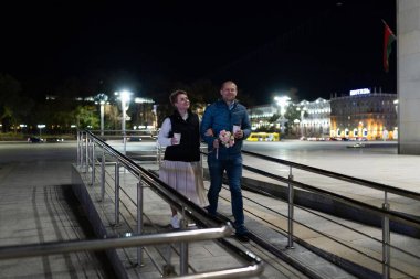 A man and woman walk side-by-side on a ramp in a city at night. They are both holding cups of coffee and the man is also carrying a bouquet of flowers. clipart