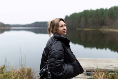 Genç bir kadın sakin bir gölün kenarında duruyor, sıcak bir ceket giymiş yeşil ağaçların ve durgun suların huzurlu çevresine dikkatlice bakıyor..