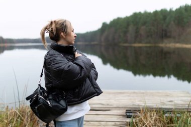 Bir kadın sakin bir gölün kenarında duruyor, kıyıda ağaçların oluşturduğu suya bakıyor. Sıcak siyah bir ceket giyiyor ve kollarını sıkı sıkı tutuyor, sakin atmosferi kucaklıyor..