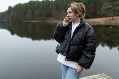 Siyah kabarık ceketli bir kadın ormandaki bir gölün yakınındaki ahşap bir rıhtımda telefonuyla konuşuyor..