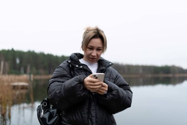 Kısa sarı saçlı bir kadın gölün kenarında duruyor. Siyah bir kabarık ceket giyiyor ve telefonuna bakıyor. Hava bulutlu ve göl sakin..