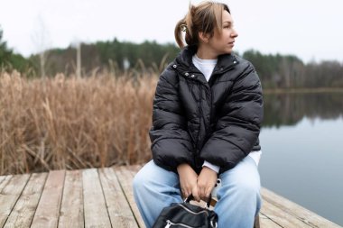 Siyah bir kabarık ceket ve kot pantolon giyen bir kadın ahşap iskelede oturuyor. O bir göle bakıyor, elleri kucağındaki siyah bir torba üzerinde dinleniyor. Arkasında uzun bir çim tarlası var..