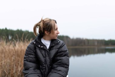 Atkuyruklu açık kahverengi saçlı bir kadın siyah bir kabarık ceket giyiyor ve sakin bir gölün üzerinden sağa bakıyor. Arka planda ağaçlar ve uzun otlar var..