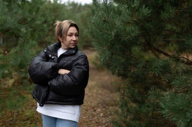 A woman wearing a cozy black puffer jacket stands with her arms crossed, gazing thoughtfully as she enjoys a tranquil moment among tall pine trees in the forest. clipart