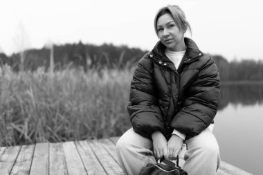 The woman sits on a wooden dock beside a serene lake, surrounded by tall grass and trees, wearing a warm puffy jacket and relaxed pants, lost in thought. clipart