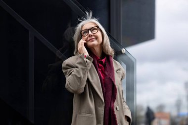 Yaşlı bir iş kadını ofisinin dışında duruyor ve bir telefon görüşmesi yapıyor. Kendine güveni tam ve düşünceli görünüyor. Şık bir ceket giymiş..