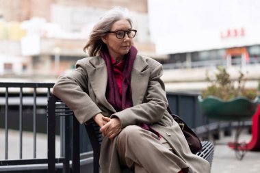 An elderly Caucasian woman dressed in a stylish outfit sits calmly on a bench, wearing glasses and a scarf, taking in her surroundings in a lively urban environment.