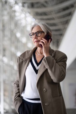 Gümüş saçlı ve gözlüklü yaşlı bir kadın telefon konuşmasına odaklanmış, kendine güvenini ve tarzını yansıtan şık bir kıyafet giymiş, çağdaş bir ortamda duruyor..