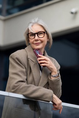 Orta yaşlı beyaz bir iş kadını, bej bir ceket giymiş, elinde kalemle bir balkonda duruyor. Çağdaş bir ofis ortamında özgüven ve duruş sergiliyor..