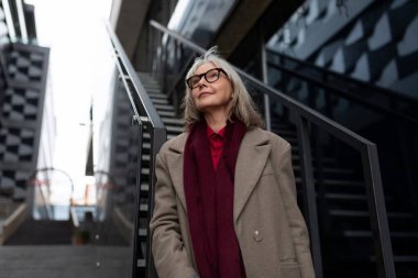 Gözlüklü ve ceketli yaşlı bir kadın modern binaların arasındaki basamaklarda dikiliyor, düşünceli bir şekilde uzaklara bakıyor, etrafındaki atmosferi kucaklıyor..