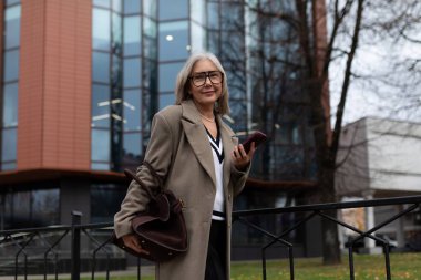Yaşlı bir kadın modern cam bir binanın yanında duruyor, elinde akıllı bir telefon ve bir çanta, bir sohbetin içinde. Hava bulutlu ve serin görünüyor..