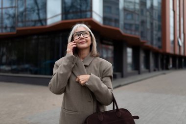 A senior businesswoman stands outside a contemporary office building, engaged in a phone conversation. She carries a stylish bag and adjusts her coat while smiling. clipart