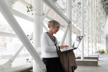 Orta yaşlı bir iş kadını, elinde palto tutarken dizüstü bilgisayarına odaklanmış, çağdaş bir ofiste yapısal bir sütunun yanında duruyor. Doğal ışık boşluğu dolduruyor ve konsantrasyonunu arttırıyor..