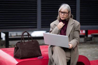 Üst düzey bir iş kadını dışarıda şık bir bankta oturuyor, dizüstü bilgisayarına odaklanmış. Şık gözlükler takıyor ve yanında dinlenen bir el çantası olan açık renk bir palto giyiyor..