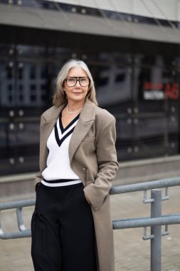 Modaya uygun yaşlı bir kadın bir korkuluğun yanında gururla durur, şık bir kıyafet giyer V yaka bir süveter ve uzun bir palto giyer, kendine güven ve zarafet saçar..