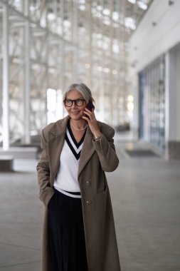 Kendinden emin yaşlı bir iş kadını, zarif bir ceket ve süveter giymiş, doğal ışıkla dolu çağdaş bir ofis alanında yürüyen bir telefon görüşmesi yapar..