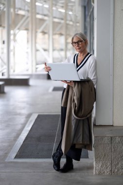 Yaşlı bir kadın lüks bir şehirde elinde dizüstü bilgisayarla bir duvara dayanıyor. Şık bir kıyafet giyiyor ve çevrenin tadını çıkarırken nişanlı ve dengeli görünüyor..