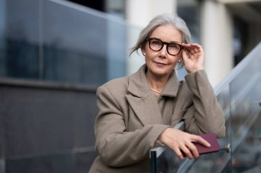 Üst düzey bir iş kadını modern bir ofisin önünde elinde akıllı bir telefon tutarken gözlüklerini ayarlıyor. Kıyafetlerine özgüven ve profesyonellik katıyor..