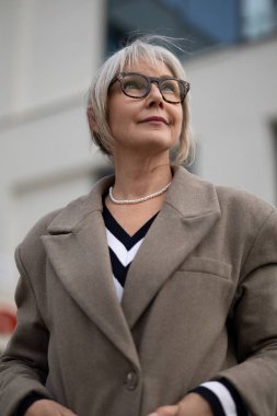 Modaya uygun yaşlı bir kadın güvenli bir şekilde dışarıda duruyor, düşünceli bir ifadeyle yukarıya bakıyor. Şık elbisesi bej bir ceket ve siyah beyaz bir kazak içeriyor. Tarzını ön plana çıkarıyor..