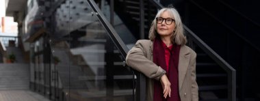 Kendinden emin, orta yaşlı, beyaz bir iş kadını elini korkuluklara uzatmış, çağdaş bir ofis binasının dışındaki kentsel bir yere dikkatlice bakıyor..