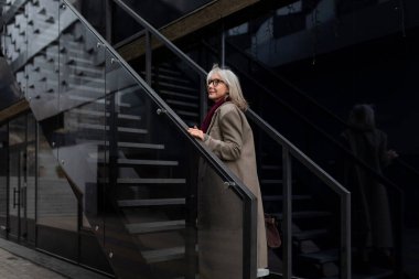Şık gri saçlı yaşlı bir kadın zarif bir şekilde giyinmiş çağdaş bir merdivene tırmanıyor ve şık, kentli bir arka plana karşı güvenini sergiliyor..