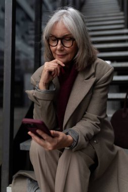 Orta yaşlı bir iş kadını şehir ortamında merdivenlere oturmuş, akıllı telefonunu dikkatle kullanıyor. Çok düşünceli görünüyor, şık bir ceket ve gözlük takmış..