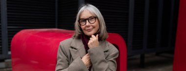 Yaşlı bir iş kadını ofisinin önünde duruyor, özgüven ve zarafet saçıyor. Şık bir palto ve gözlük takıyor, şehirde sükunetin tadını çıkarırken gülümsüyor..