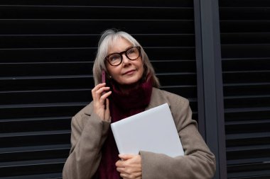 Yaşlı bir kadın şık bir binanın önünde elinde bir defterle telefonuyla sohbet ediyor. Sıkı giyinmiş ve sohbete katılmış gibi görünüyor..