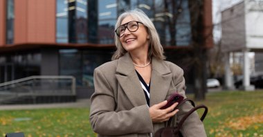 Şık bir palto giymiş yaşlı bir kadın sonbahar havasının tadını çıkarırken gülümseyerek parkta dolaşıyor. Arkaplanda çağdaş bir bina yer alıyor.