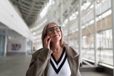 An older woman stands in a contemporary indoor space, her thoughtful expression highlighting her style. She looks up as if contemplating the surroundings, showcasing her confidence. clipart