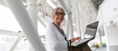 Orta yaşlı beyaz bir iş kadını, çağdaş bir ofis ortamında dizüstü bilgisayarını kullanarak gülümsüyor. Parlak alan onun profesyonel davranışlarını vurguluyor..