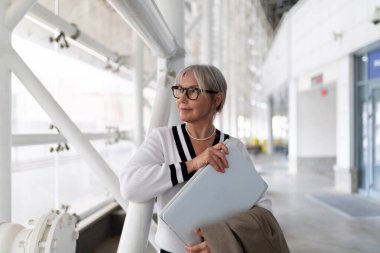 Yaşlı bir iş kadını ofisinin önünde duruyor, elinde göğsüne yakın bir laptop çantası var. Düşünceli bakıyor, profesyonel kıyafetler giyiyor, kendine güveni tam..