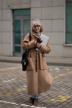 An older woman dressed in a fashionable long coat is standing outside a modern building. She holds a laptop and some notes, exuding confidence and style in a chilly environment. clipart