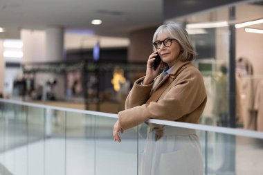 Hareketli bir alışveriş merkezinde, gözlüklü şık, olgun bir kadın bir telefon görüşmesi yapıyor, korkuluklara yaslanıyor, çağdaş şehir atmosferini gösteriyor..