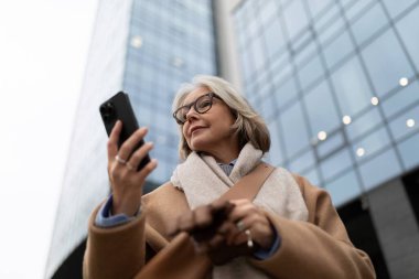 Olgun bir kadın, çağdaş bir binanın önünde duruyor, akıllı telefonuyla kayıyor. Sıcak bir palto ve eşarp giyer, şehir ortamında rahat ve nişanlı bir tavır sergiler..