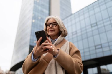 Gri saçlı, gözlüklü ve şık ceketli bir kadın bulutlu bir günde şehir mimarisinin ortasında akıllı telefonuna odaklanıyor..