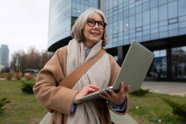 Üst düzey bir kadın, modern bir ofis binasının önünde dizüstü bilgisayarında yazarken gülümsüyor. Ayar, serin bir günde doğa ve şehir hayatının bir karışımını yansıtıyor..