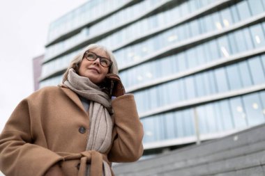 Gri saçlı ve gözlüklü bir kadın telefonla konuşuyor. Sıcak bir palto ve eşarp giyiyor. Çağdaş bir binanın dışındaki soğuk havaya hazır..