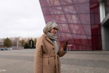 Kadın, çarpıcı bir çağdaş yapının önünde telefonuyla etkileşime girerken neşeli görünüyor. Hava serin, sonbahar ya da kış başlarını öneriyoruz..