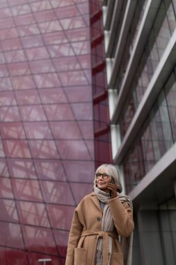 An elderly woman wearing a stylish tan coat and scarf engages in a cheerful phone call near a contemporary glass building. The urban landscape reflects modern architecture and design. clipart