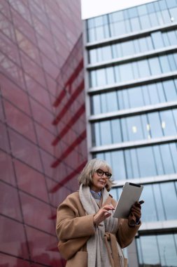 A woman with gray hair and glasses stands in an urban setting, focused on her tablet. The striking red and glass architecture surrounds her as she engages with technology. clipart