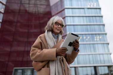 Yaşlı bir kadın dışarıda duruyor, sıcak kahverengi bir ceket ve eşarp giymiş, rahatça tablet kullanıyor. Bulutlu bir günde modern mimari yapılarla çevrilidir..