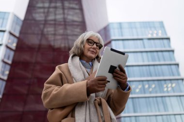 Son sınıf öğrencisi bir kadın, çağdaş binaların dışında duran bir tabletle meşgul. Yansıtıcı cam ve kalın yapı alacakaranlık çökerken çarpıcı bir zemin oluşturur..